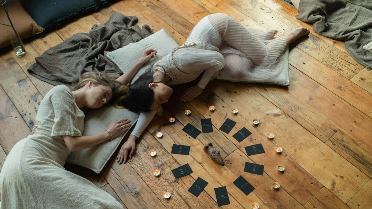Two women lying on wooden floor, surrounded by candles and tarot cards, in a spiritual setting.