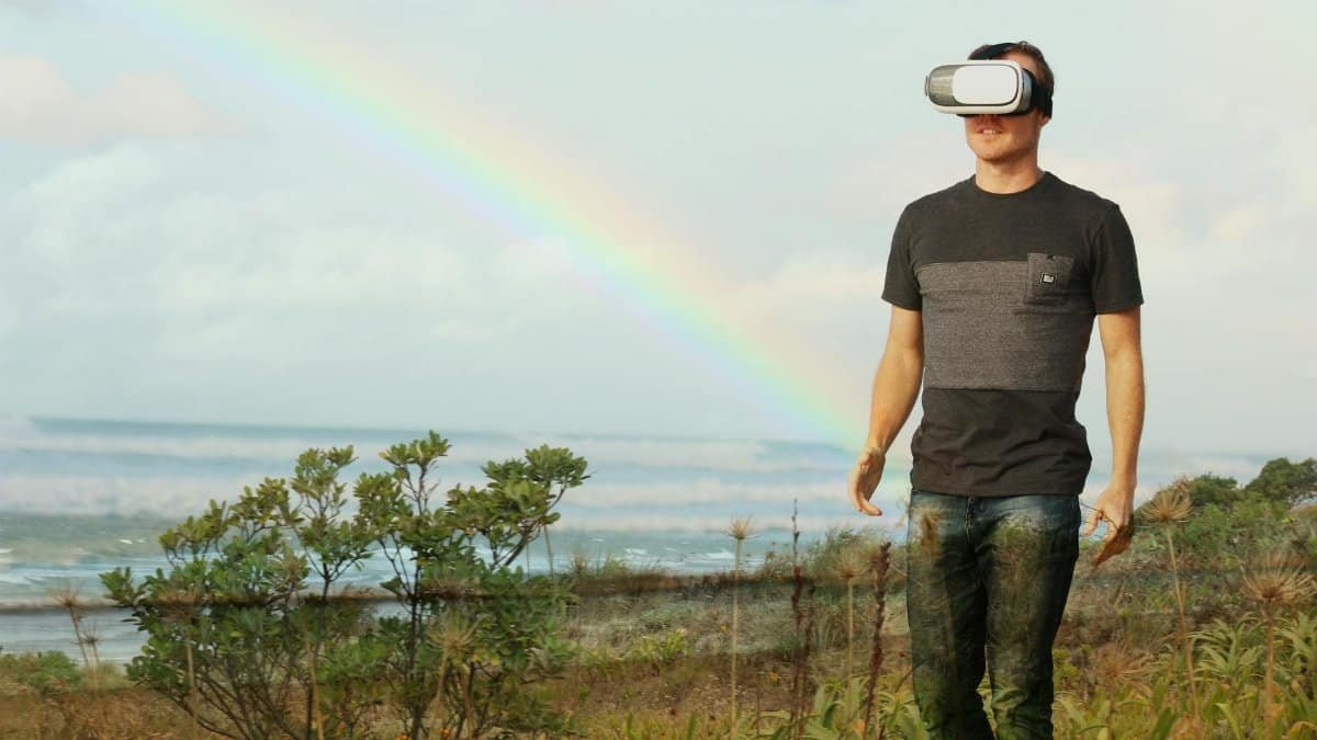 Man using VR headset walks in nature under a rainbow, showcasing modern technology outdoors.