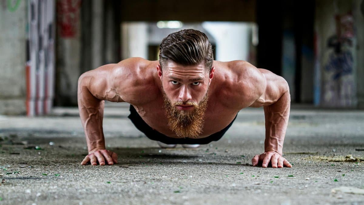 Fit shirtless man with a beard doing a push-up exercise on concrete floor.