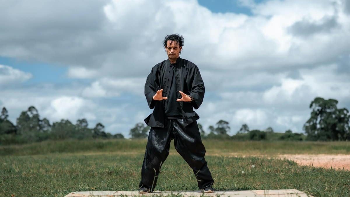 Man practicing martial arts outdoors in black uniform, performing hand gestures on a cloudy day.