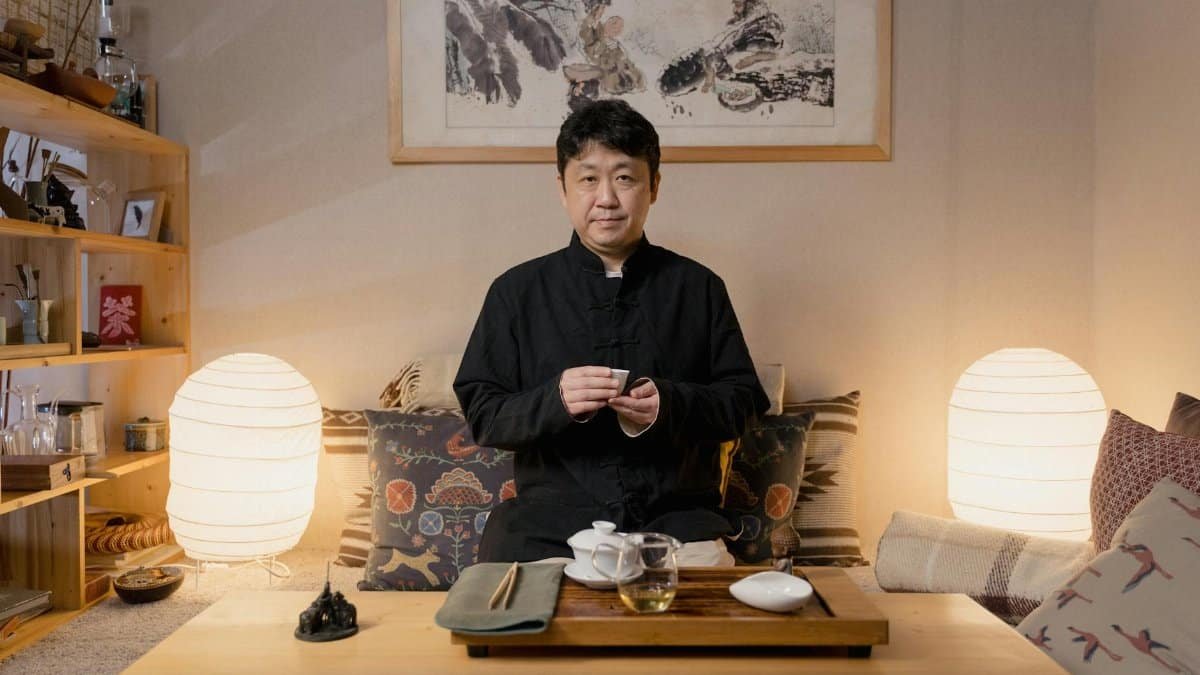 An East Asian man practicing a traditional tea ceremony in a cozy, dimly lit tea room.