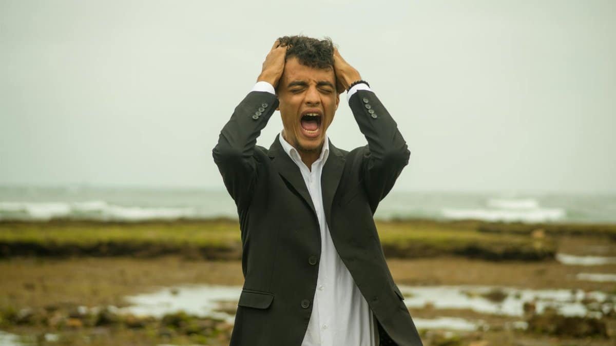 A man in a suit screams in frustration while standing on a windy beach. Emotional stress concept.