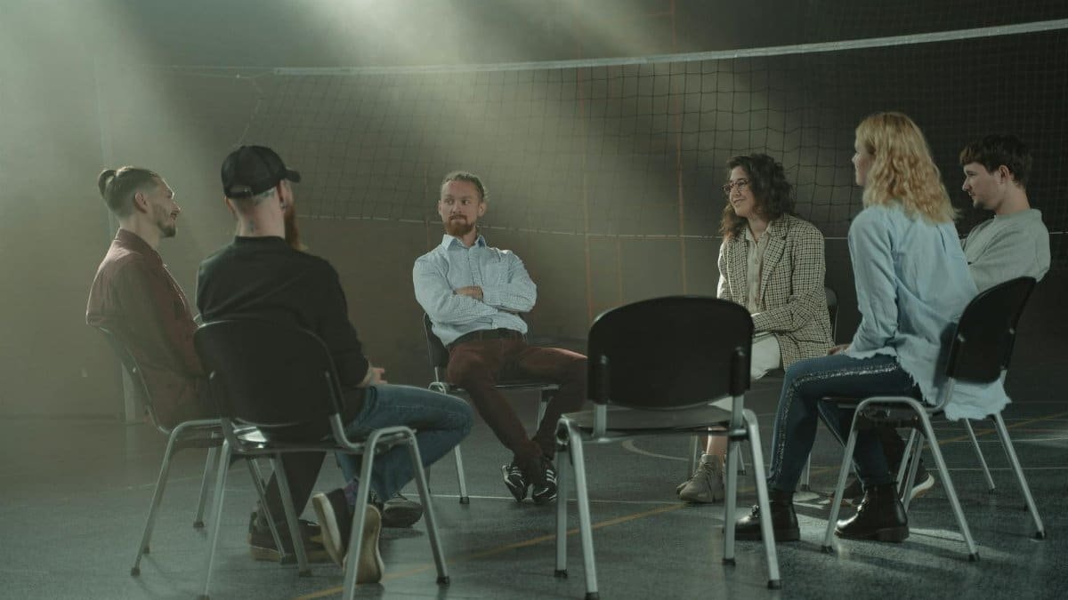 A group therapy session with six adults seated in a circle, discussing support and mental health.