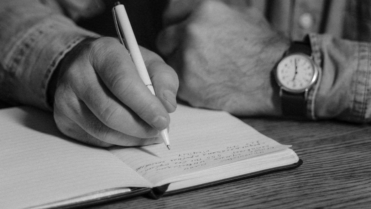 Black-and-white close-up of person writing in a notebook, emphasizing handwriting and detail.