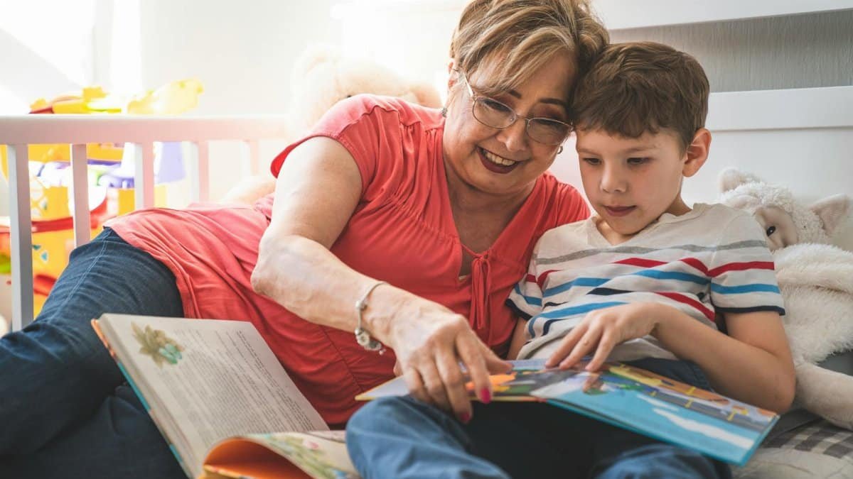 Elderly woman reading a book with her grandson on a cozy bed in a sunlit room.