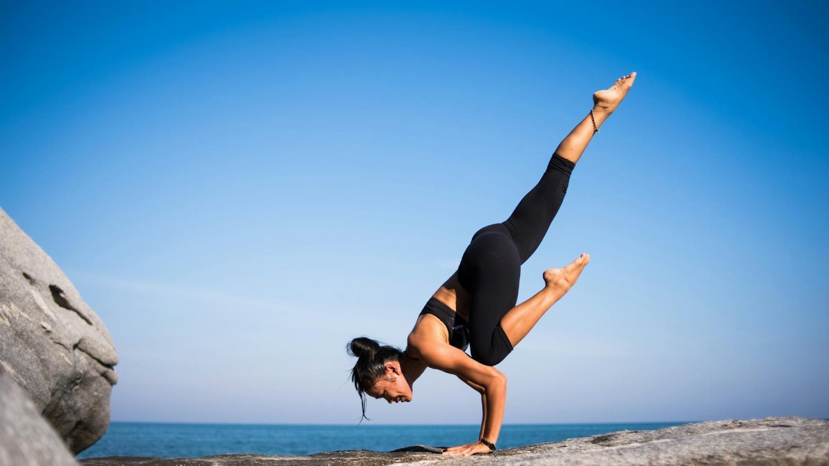Woman performing a yoga pose on a rocky beach with ocean view and clear blue sky.