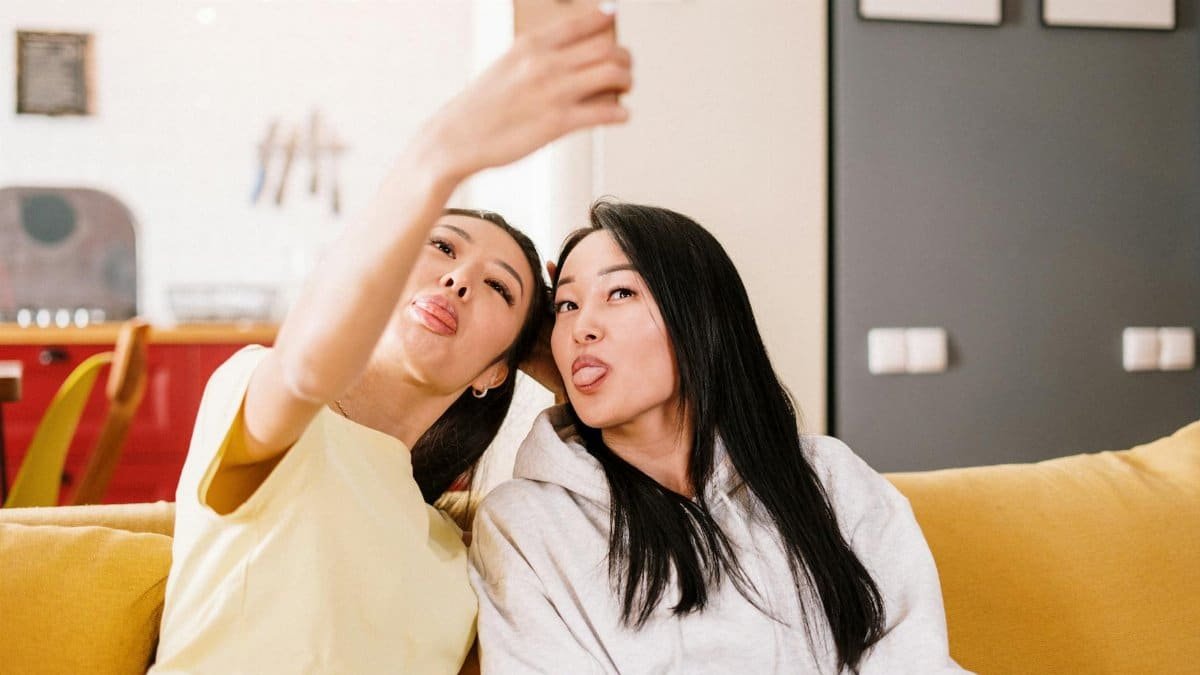 Two friends make funny faces while taking a selfie on a cozy sofa at home.