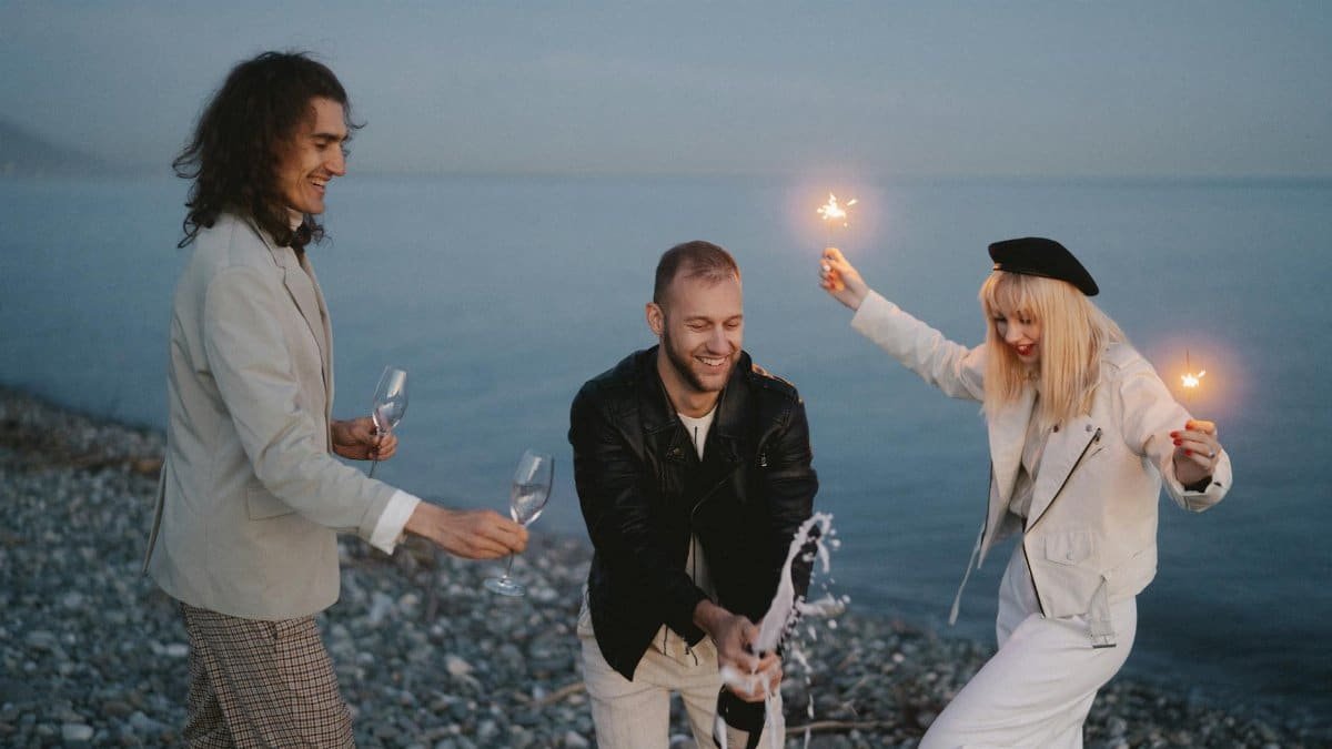A group of friends joyfully celebrate with sparklers and champagne by the ocean at twilight.