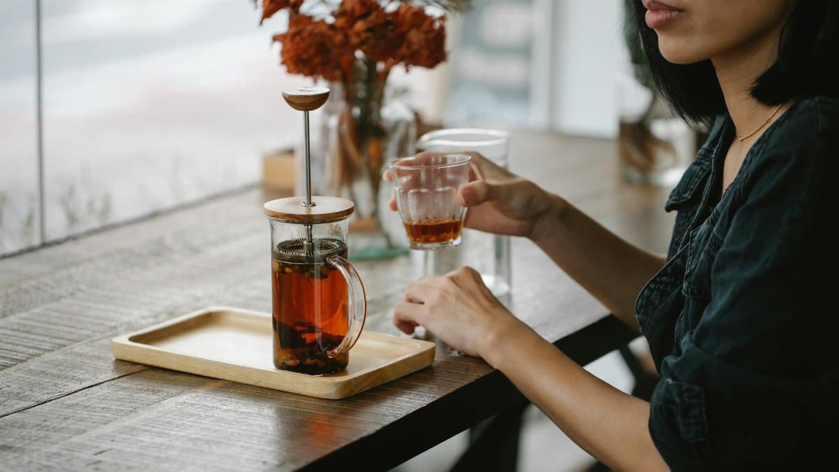 A woman enjoys a calming tea break with a French press on a wooden table in a cozy café.