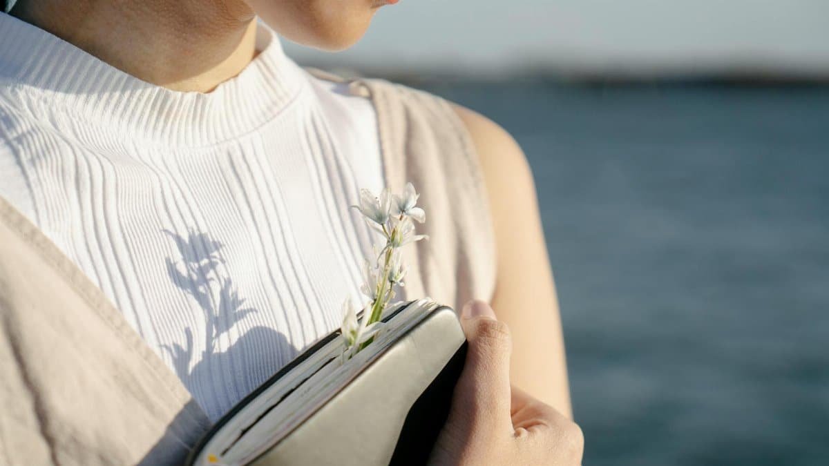 Crop unrecognizable gentle female with blossoming flower in diary against river on sunny day