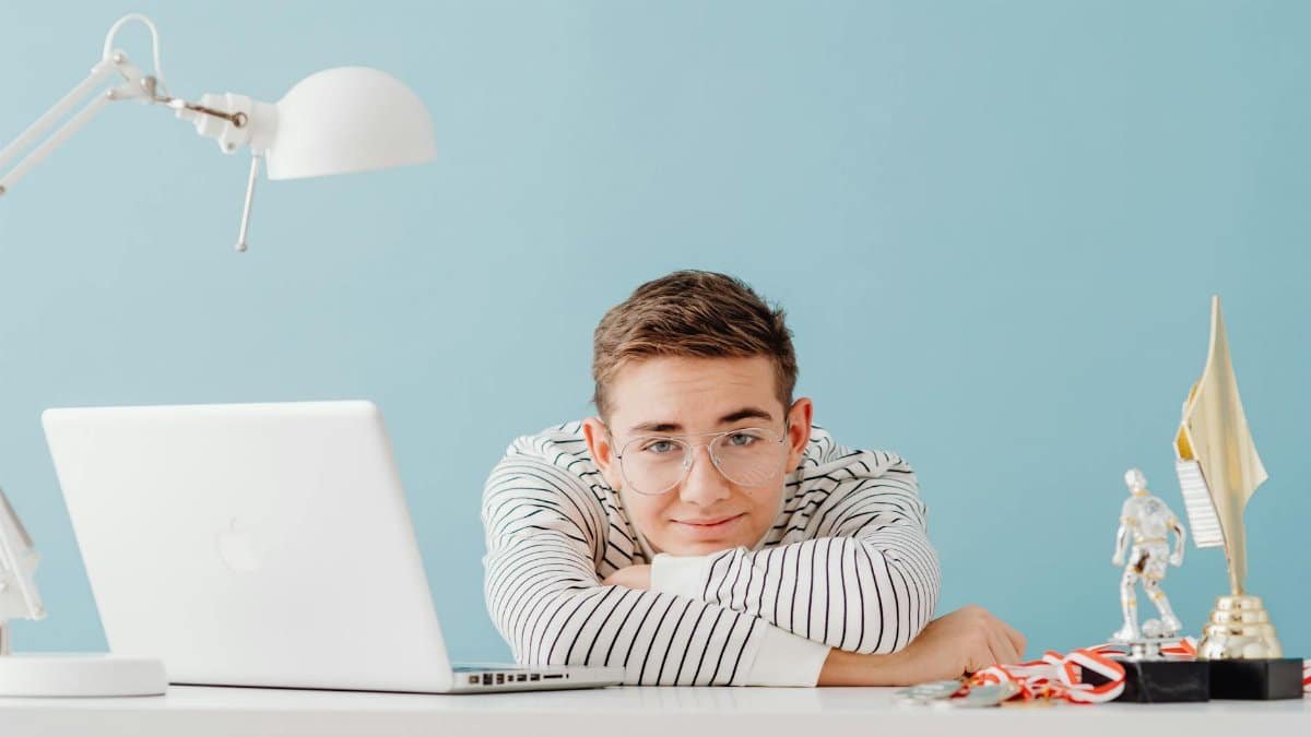 Young man with glasses leaning on a desk with a laptop and trophies, against a teal background.