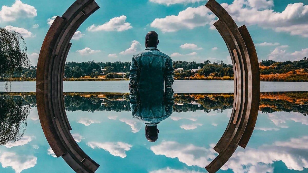A man in a denim jacket sits by abstract sculpture reflecting on the water under a blue sky.