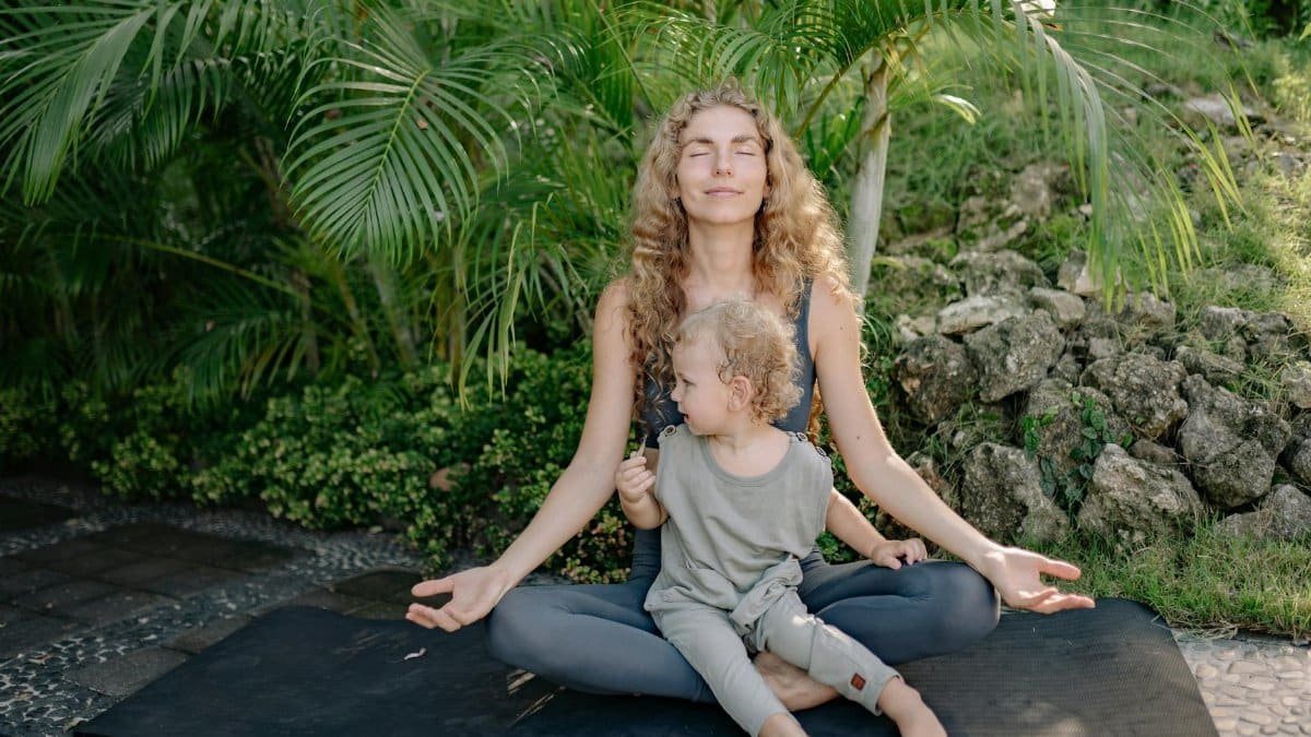 Full body happy relaxed mother sitting with eyes closed in lotus pose with cute boy on laps while practicing yoga in tropical green park