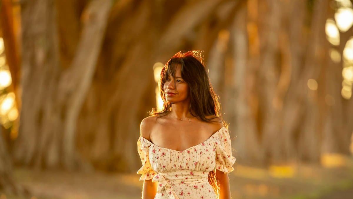 A woman in a floral dress stands on a sunlit forest path, embracing nature's tranquility.