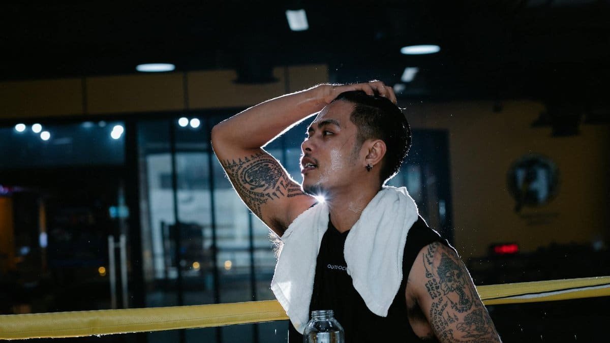 Side view of tired tattooed Latin American male in sportswear with white towel around neck standing in gym with bottle of water during workout
