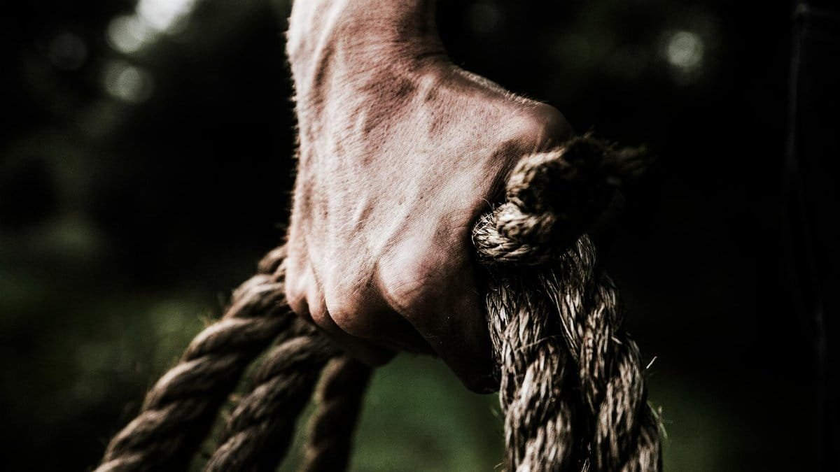 A close-up image shows a strong, firm hand gripping a thick, rough rope, showcasing strength and determination in an outdoor setting.