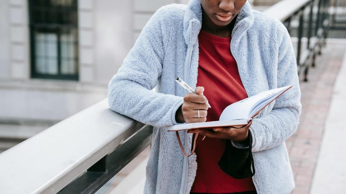 Crop concentrated ethnic female student in casual apparel watching diary near fence in town