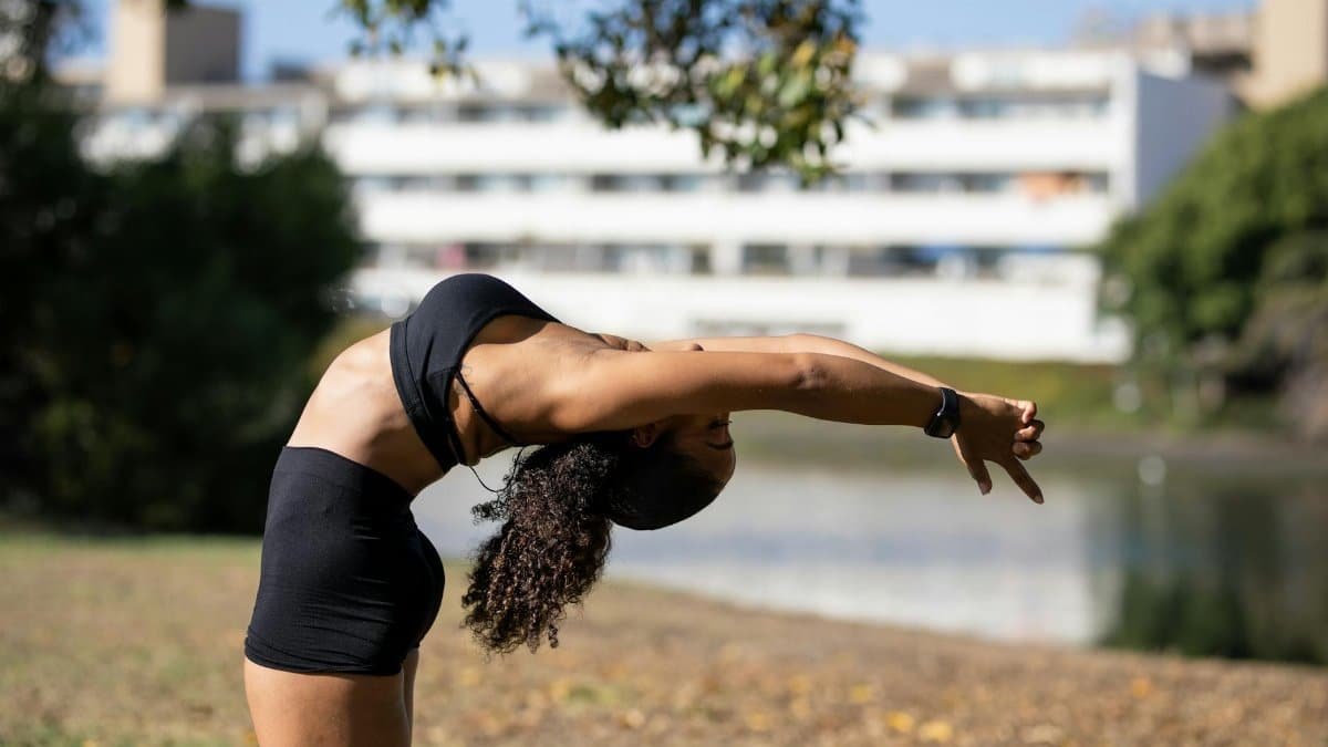 A woman exercises outdoors, performing stretching poses in a sunny urban park with blurred buildings in the background.