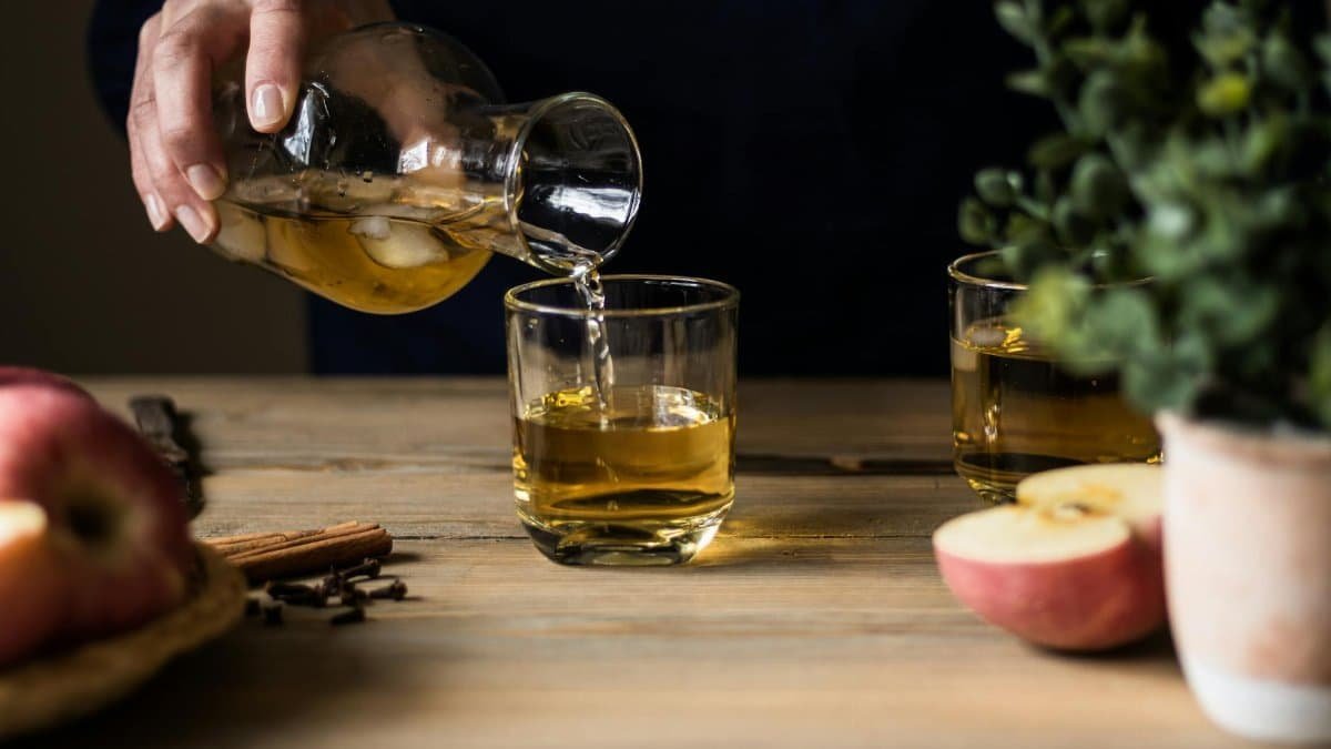 Close-up of someone pouring apple cider into glasses on a wooden table with apples and spices.
