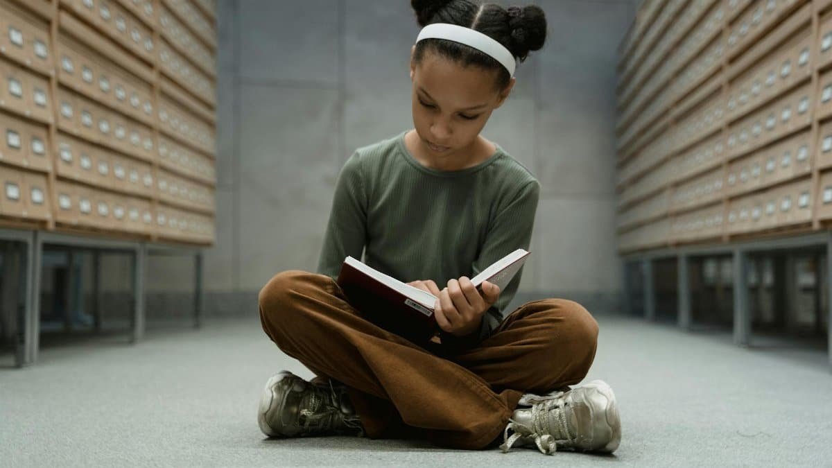 A young girl sitting cross-legged, reading a book in a library with card catalogs.