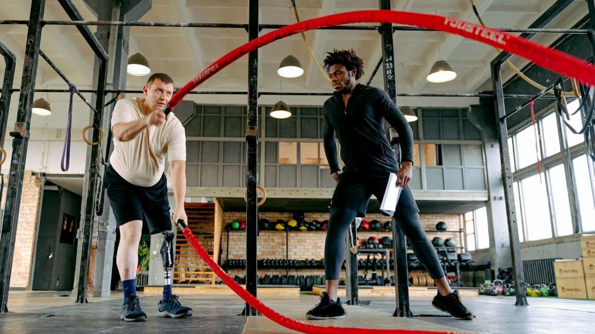 An adult male with a prosthetic leg exercises with ropes, guided by a personal trainer in a gym.