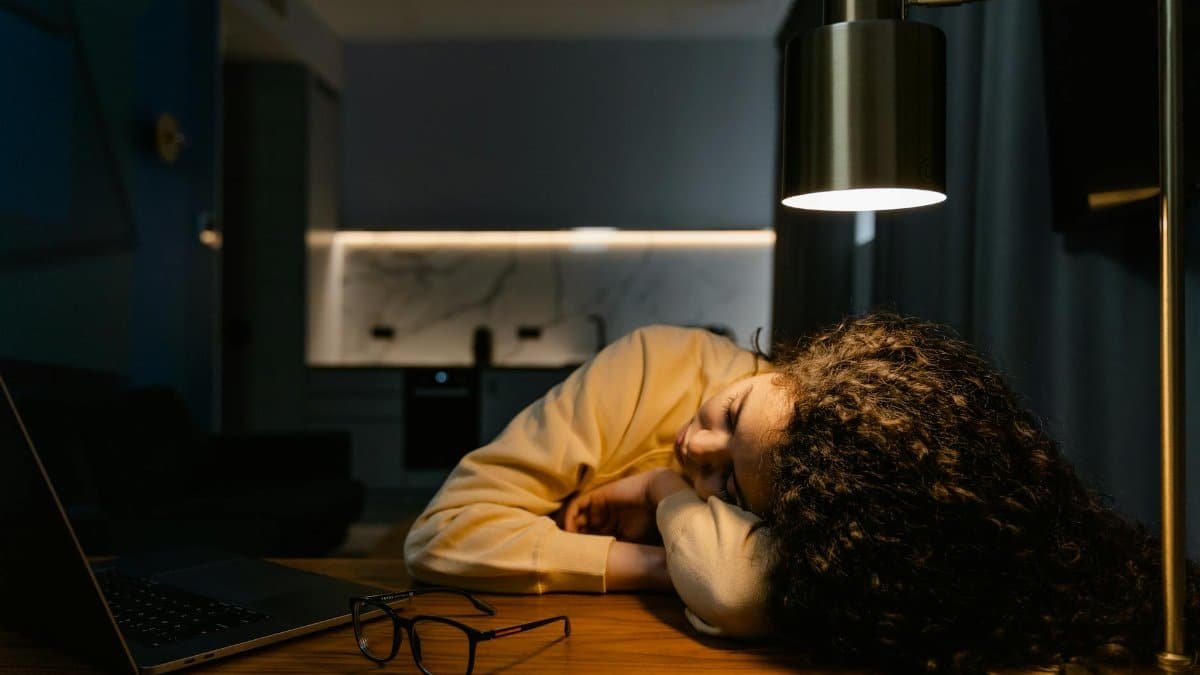 Exhausted woman sleeps at her desk under a lamp with a laptop beside her.