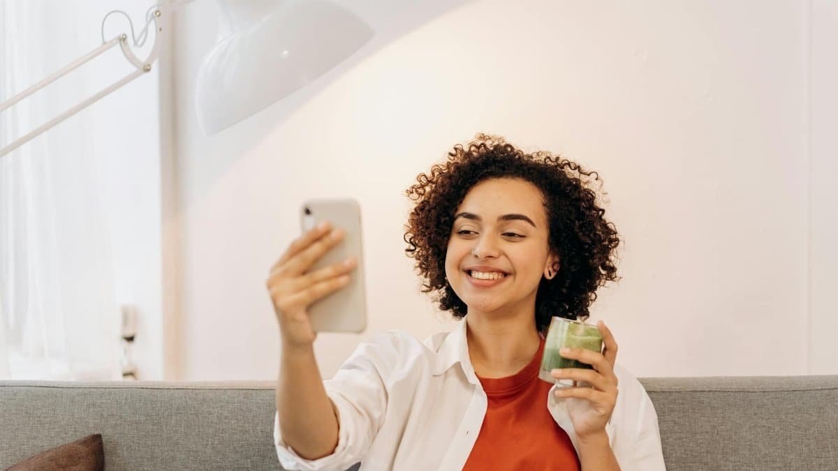 Woman sitting indoors, taking a selfie while holding a green smoothie, promoting healthy lifestyle.