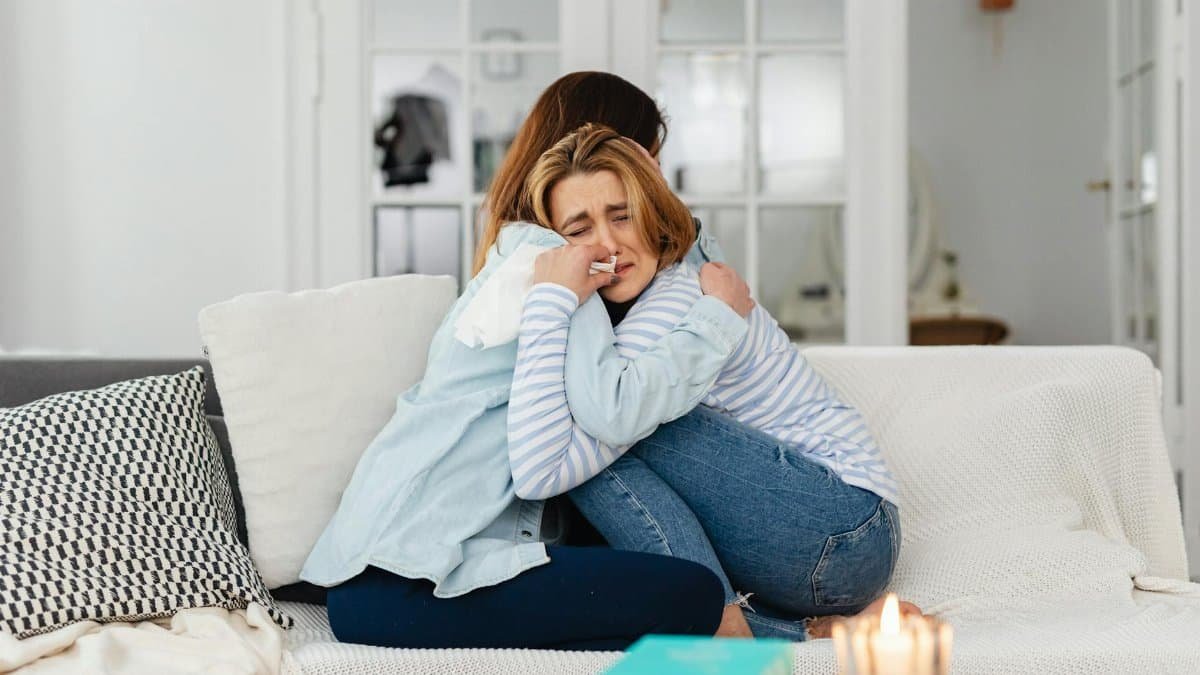 Two women embracing on a sofa, providing comfort and support in a warm, well-lit living room.