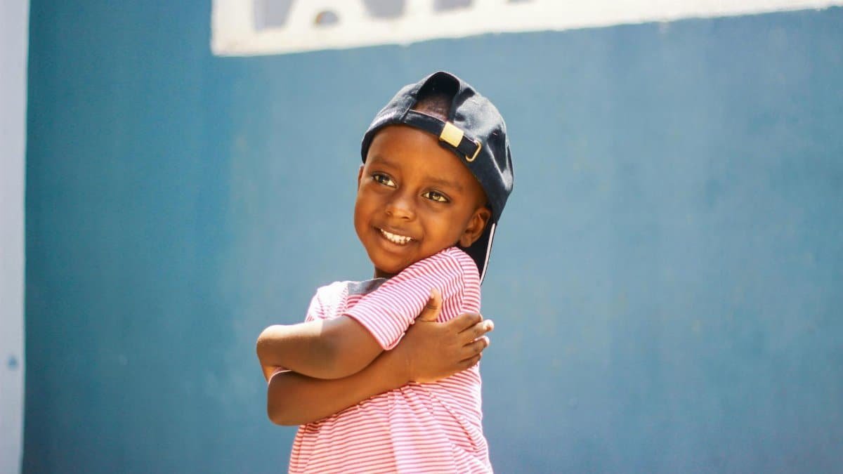 Adorable child in a striped shirt and cap smiling warmly while hugging self. Joyful and carefree moment.