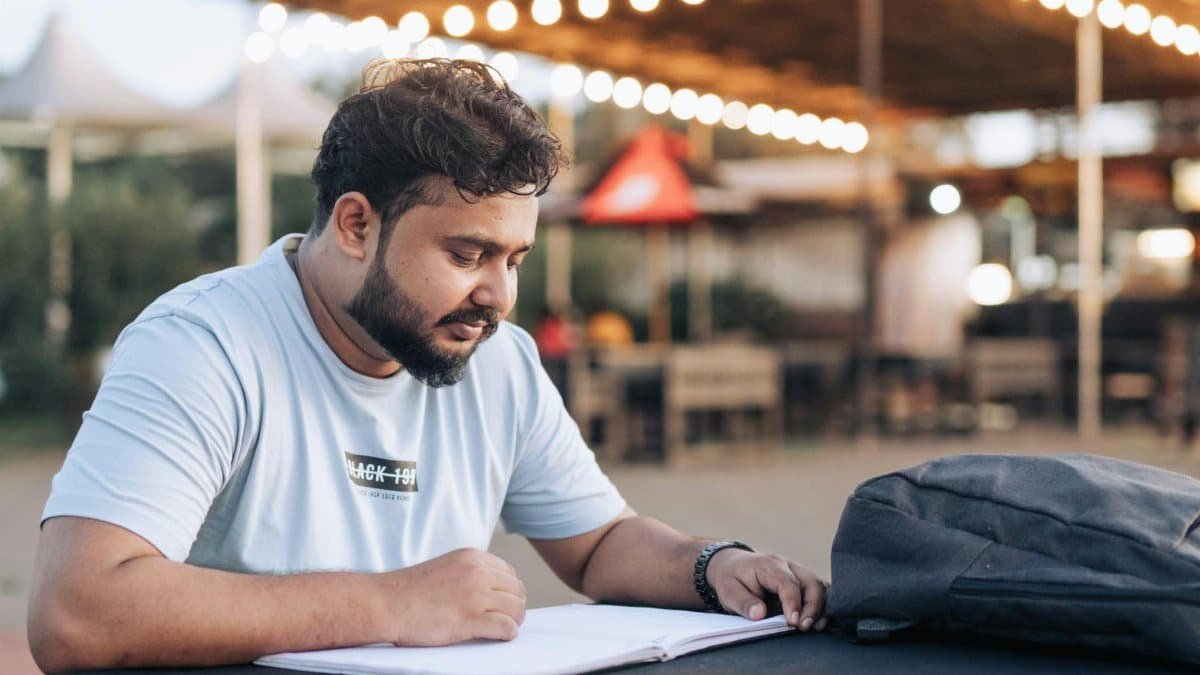 Man studying at an outdoor cafe table with a notebook and backpack in the evening.