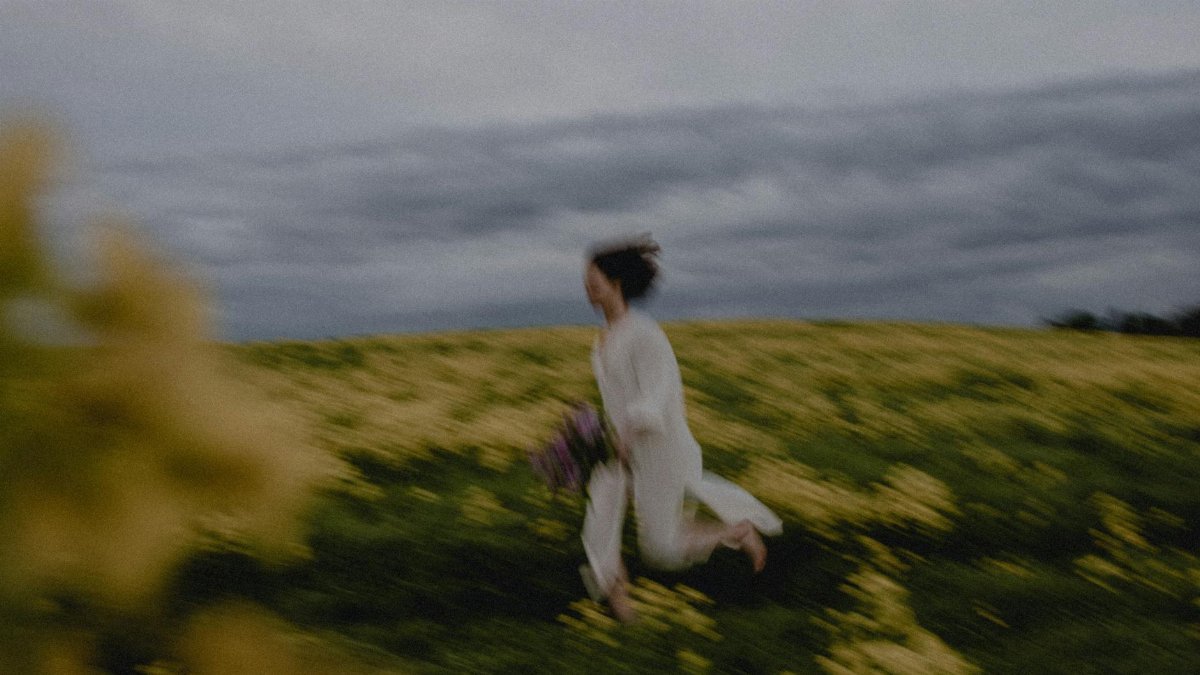 Artistic blur of a woman running, capturing movement amidst a field of yellow wildflowers under cloudy skies.
