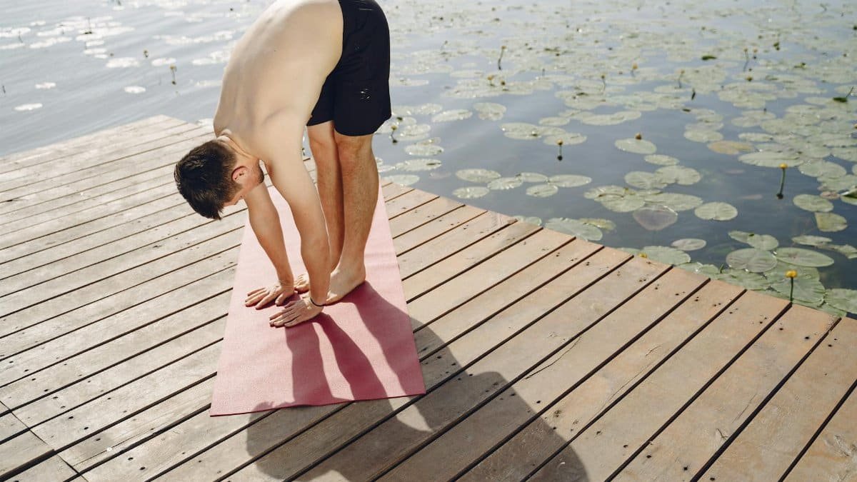 Shirtless man performing yoga on a wooden pier by a lake amidst nature, promoting relaxation and mindfulness.