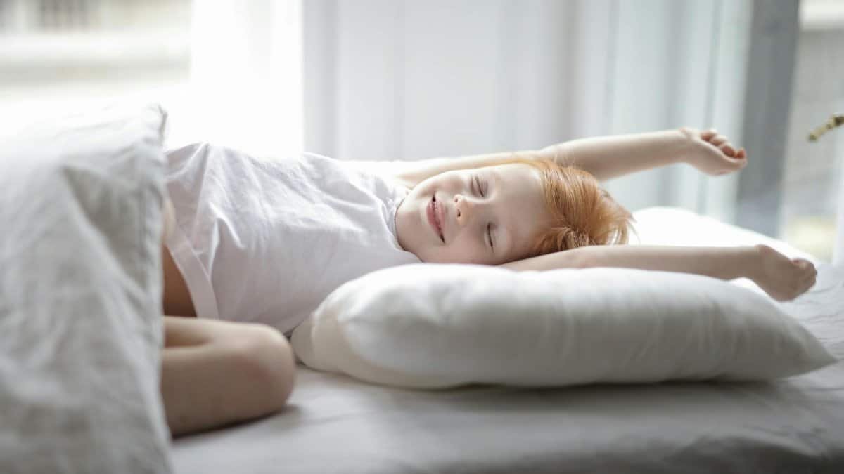 Little adorable child with red hair lying in bed with white linen while trying waking up early morning stretching hands after sleep in light cozy bedroom at home