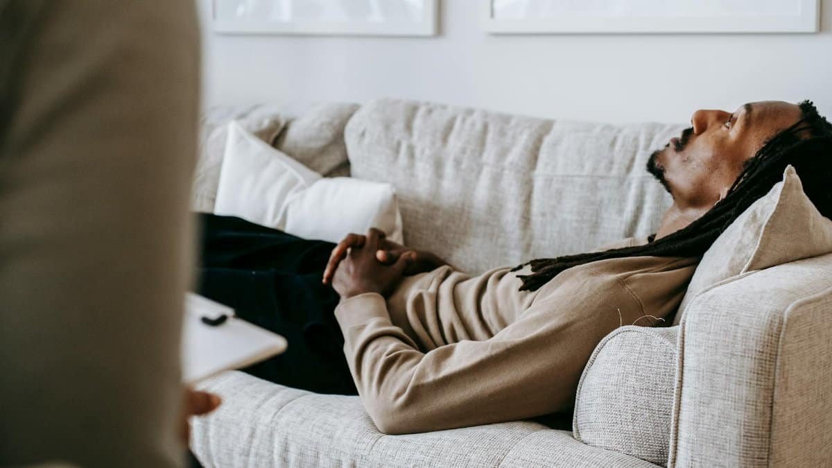 Man in therapy session lying on sofa, conversing with therapist in a relaxed indoor setting.
