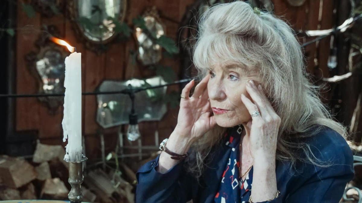 A thoughtful senior woman sits by a candle in an intimate, mirror-lined interior setting.