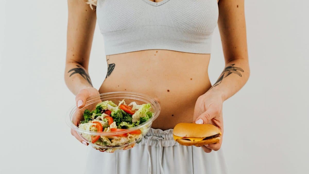 A woman in a sports bra holding a salad and cheeseburger, representing healthy vs junk food choices.