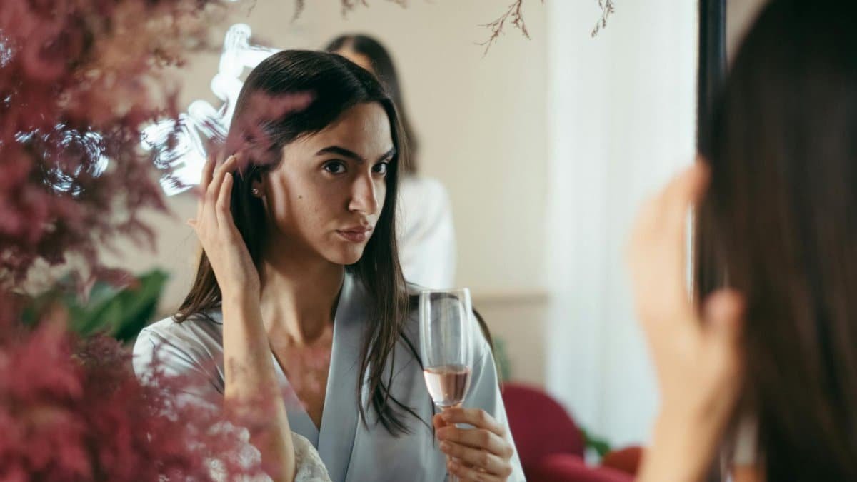 A woman in a robe holding a glass of rose champagne while looking in the mirror.