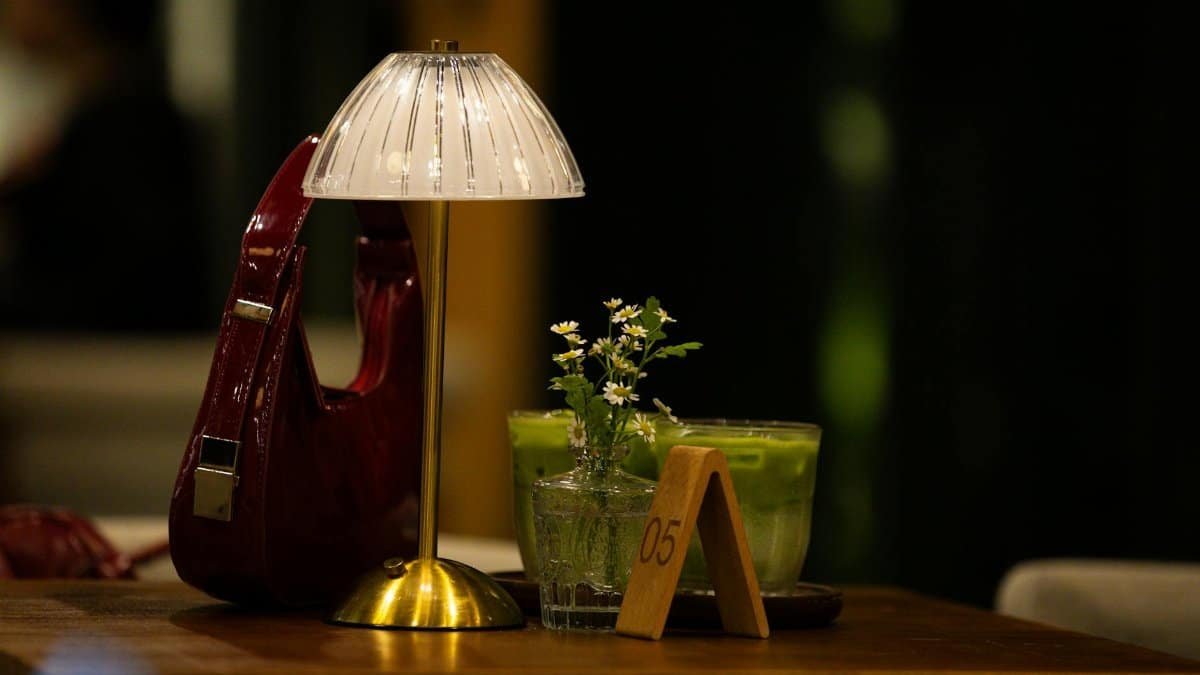 A warm cafe table setting with a lamp, handbag, green beverage, and flowers.