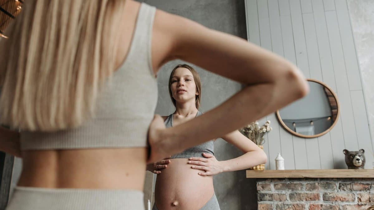 A pregnant woman practicing gentle exercise indoors, reflected in a mirror, in casual sportswear.