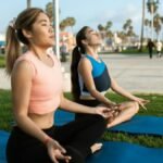 Two women meditating on yoga mats in a sunny park setting, promoting healthy living and relaxation.
