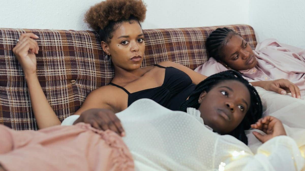 Three black women relaxing on a bed in a cozy indoor setting, reflecting a moment of calm and companionship.