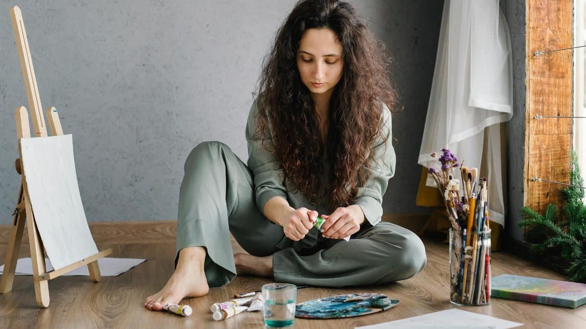 A young woman sitting on the floor in an art studio with paints and brushes, immersed in her creative work.
