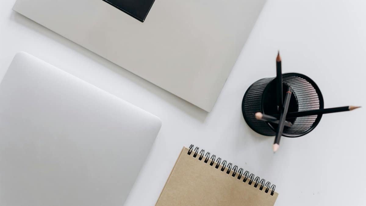 Top view of a minimalist workspace featuring a laptop, notepad, and stationery on a white surface.