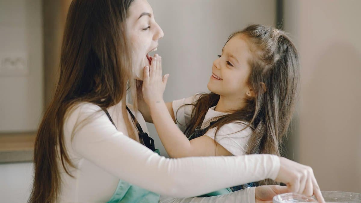 Side view of young woman and little girl in aprons playing and enjoying each other while kneading dough together on modern kitchen