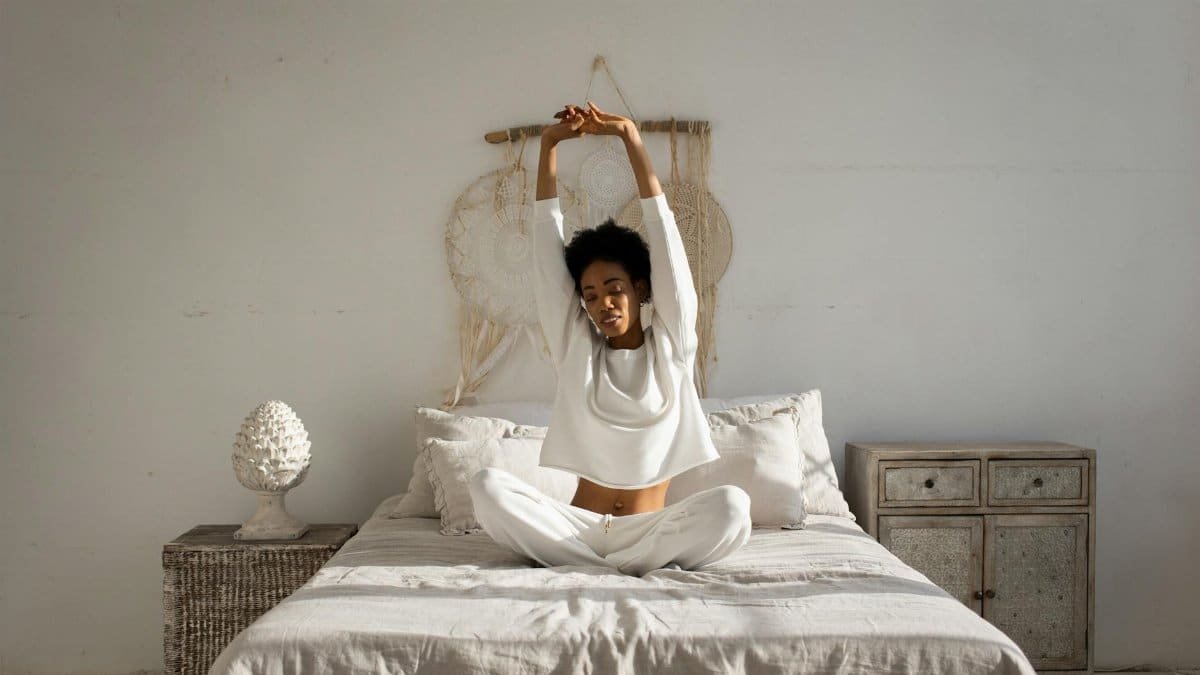 African American woman in white sitting on bed, stretching arms in a cozy minimalist bedroom with natural light.