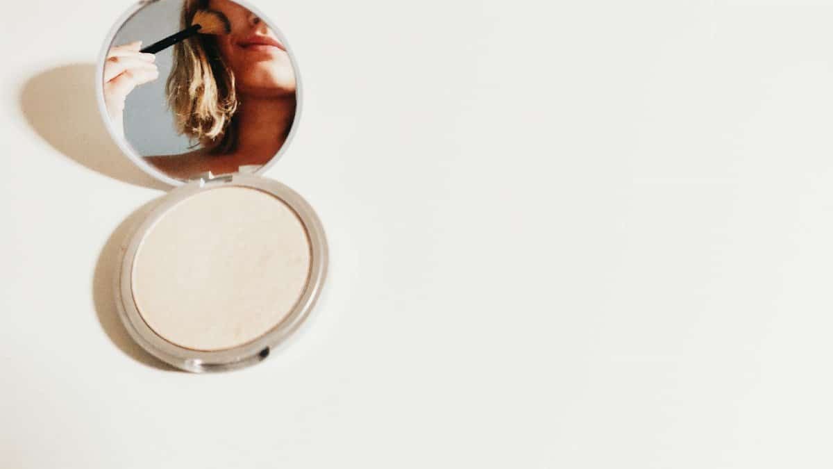 Close-up of a woman using makeup in a compact mirror, set against a bright background.