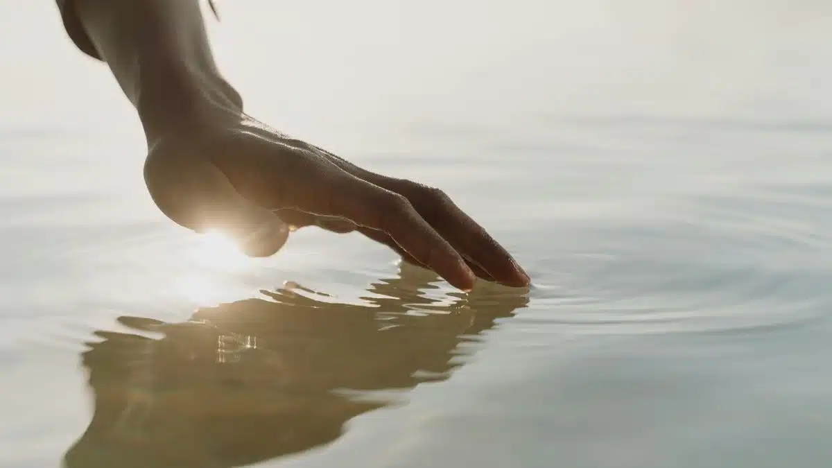 A tranquil scene of a hand gently touching the water, creating ripples with gentle sunlight reflection.