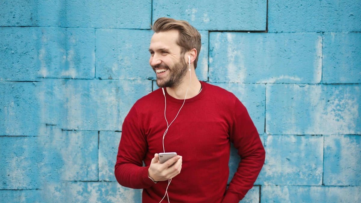 A joyful man in a red sweater smiles while listening to music on his smartphone against a blue brick wall.