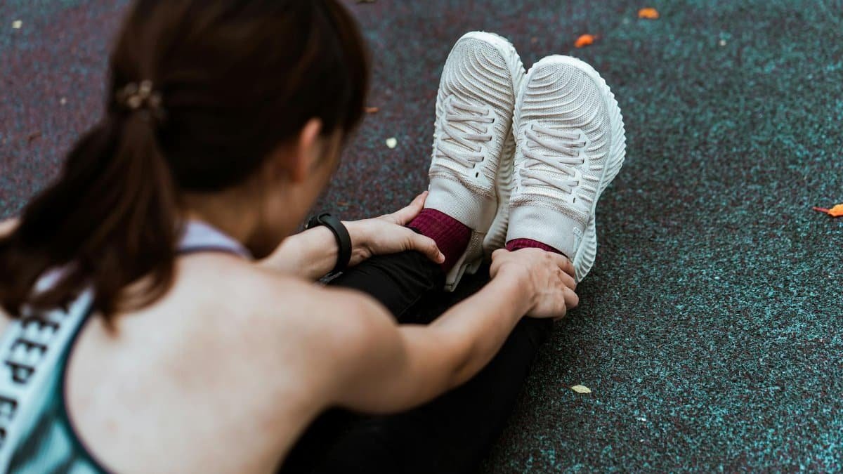 Top view of crop unrecognizable female athlete in sports clothes and sneakers doing forward bend while sitting on asphalt footpath with shabby surface