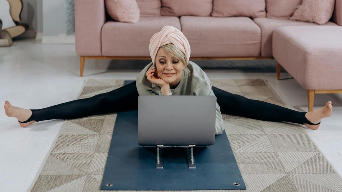 A senior woman enjoys a relaxing yoga session indoors, stretching while using her laptop.